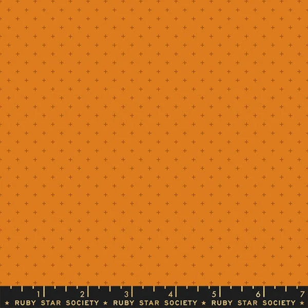 A close-up fabric or material swatch showing a warm orange background with a repeating small dark plus-sign pattern and a ruler edge along the bottom labeled Ruby Star Society.