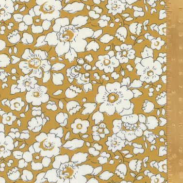 A close-up of a floral fabric pattern with white flowers and green stems on a mustard yellow background, shown with a wooden ruler along the right edge for scale.